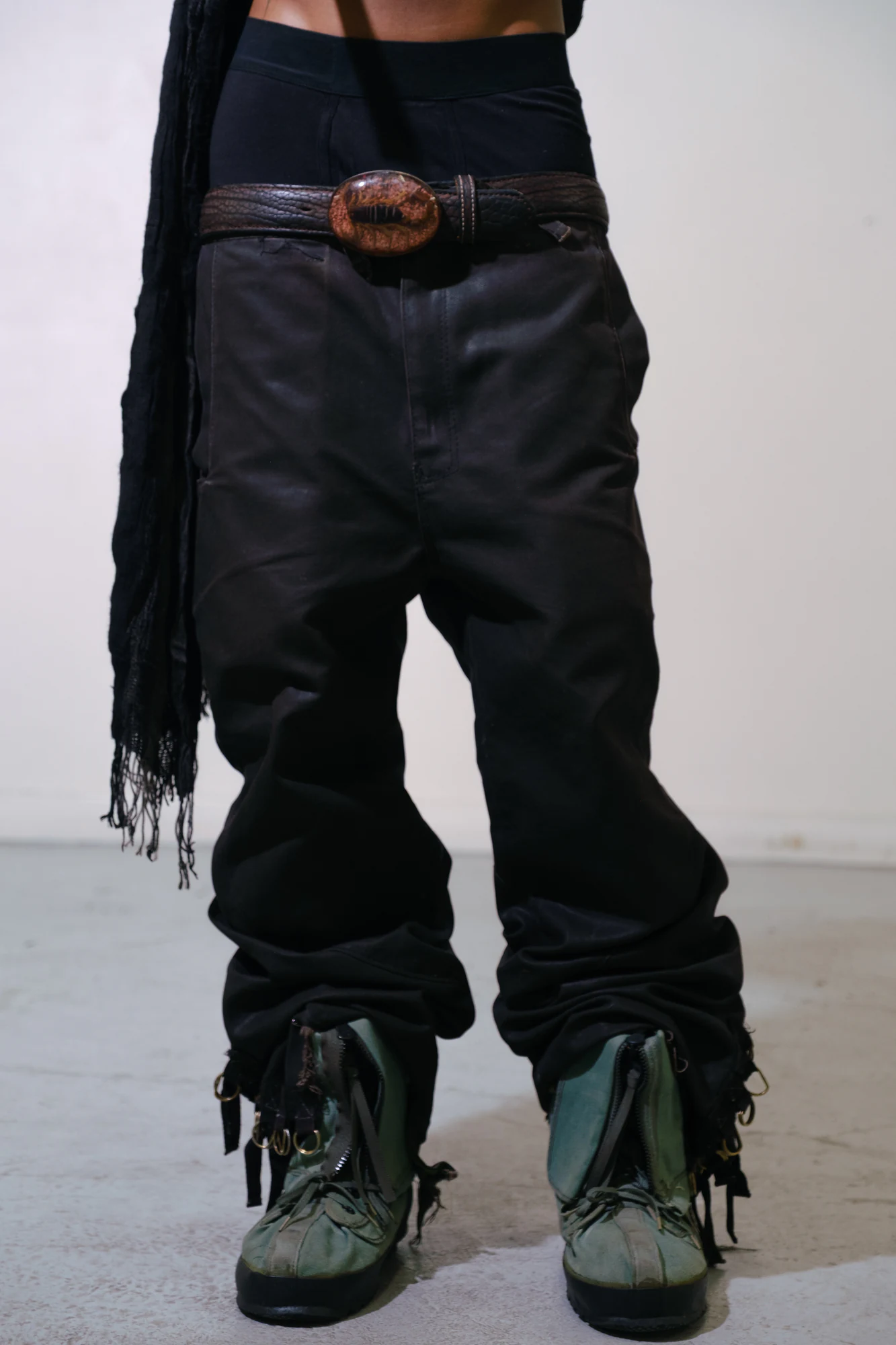 Close-up of waxed black pants, a leather belt, and strapped footwear.
